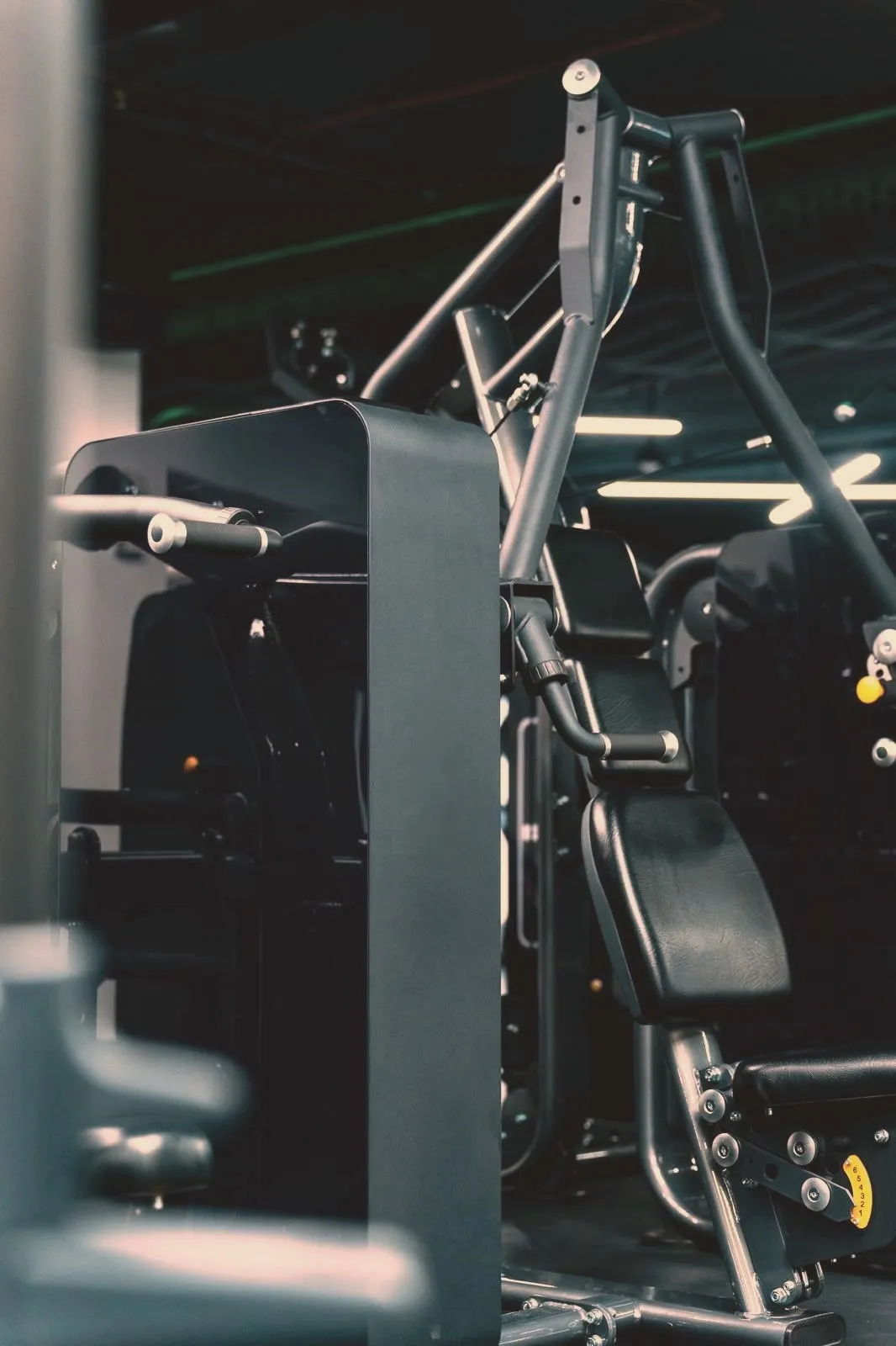A chest-press machine close and low: the dark shroud over its weight stack at the left, the seat and press arms beyond it, chrome handles and the ceiling’s light bars catching in the gloom.