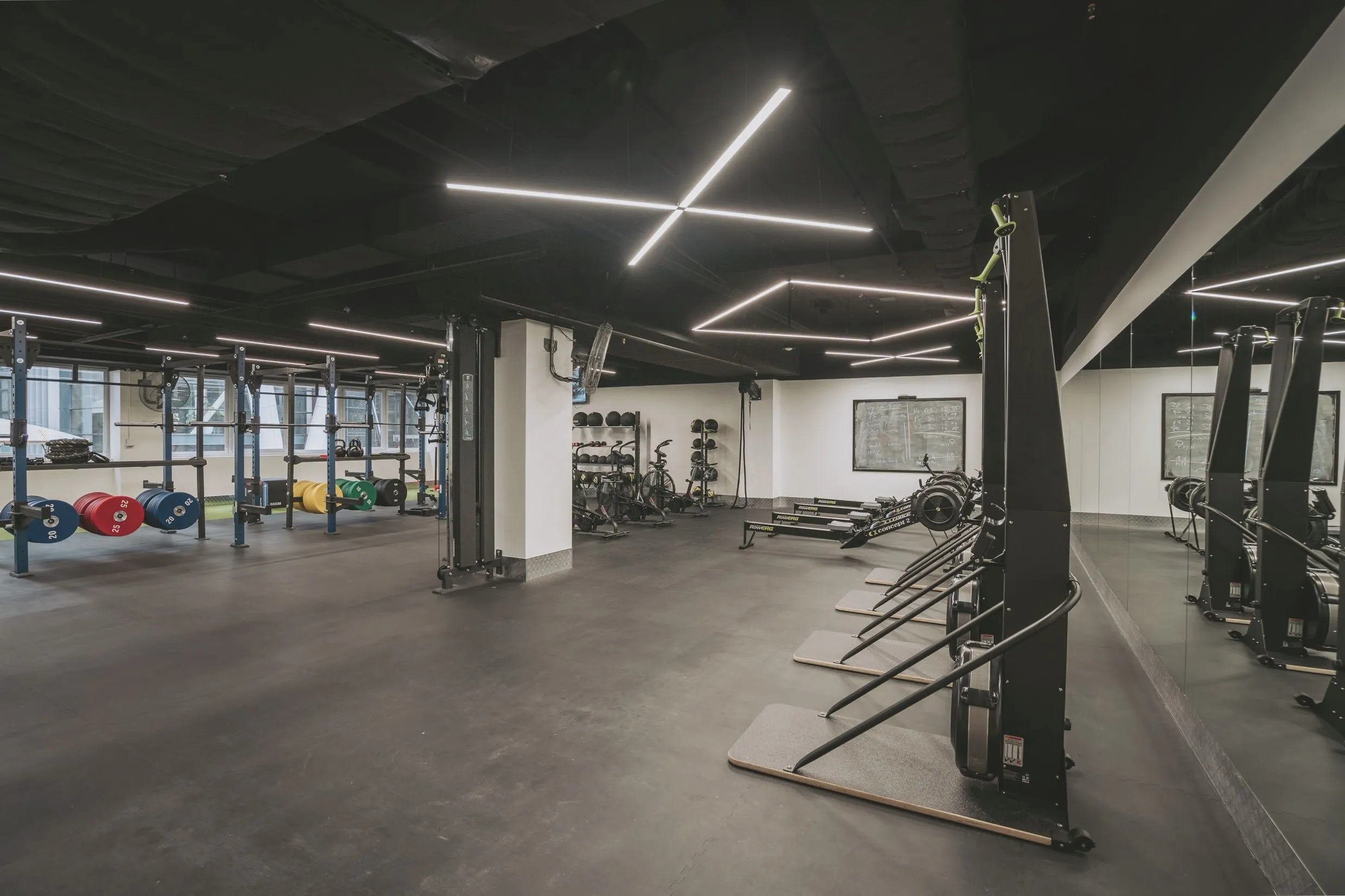The performance zone: racks and bumper plates beside a strip of turf, with ski and rowing ergometers along the mirrored wall and a chalkboard of the day’s work.