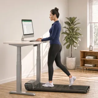 A small walking pad under a standing desk, in use: a person walks on it whilst working at their desk